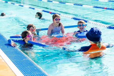 Swim instructor in pool talking to four students who are standing on a blue and red pontoon 